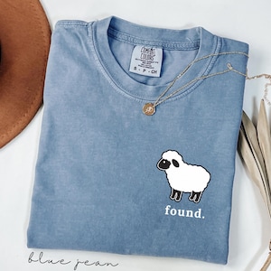 May include: A blue jean colored t-shirt with a crew neckline and a Comfort Colors brand label. The shirt features a graphic of a white sheep with a black face and legs on the lower right side, with the lowercase white text found. printed below.