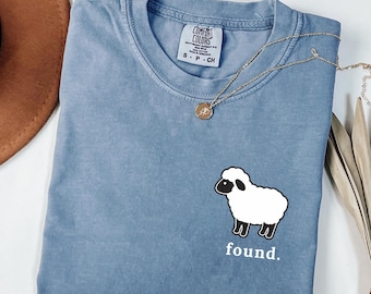 May include: A blue jean colored t-shirt with a crew neckline and a Comfort Colors brand label. The shirt features a graphic of a white sheep with a black face and legs on the lower right side, with the lowercase white text found. printed below.