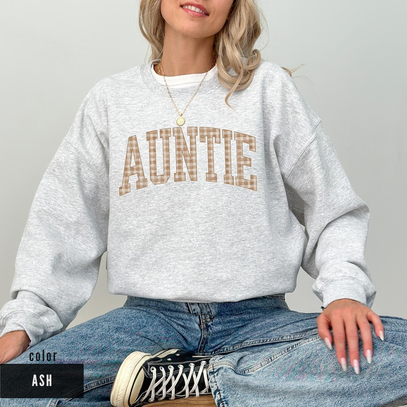 May include: A light gray sweatshirt with the word "AUNTIE" in a plaid design across the chest. The sweatshirt is a crew neck style and is shown in the color "ASH". The model is wearing blue jeans and black and white sneakers.