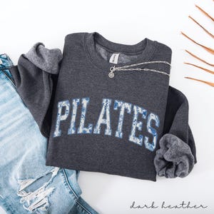 May include: Dark heather gray sweatshirt with the word "PILATES" in white letters with a blue floral design. A silver necklace with a circular pendant rests on the sweatshirt. The sweatshirt is paired with light blue jeans.