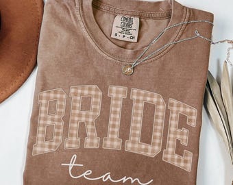 May include: Espresso-colored t-shirt with the words "BRIDE team" in a plaid design. The shirt is from Comfort Colors and has a small, round pendant necklace. A brown hat is visible in the upper left corner.