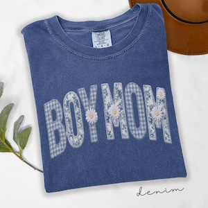 Peut inclure: T-shirt bleu denim avec l'inscription "BOY MOM" en motif floral et vichy, ornée de petites marguerites blanches. Le mot "denim" est écrit en cursive en bas du t-shirt. Marque Comfort Colors.