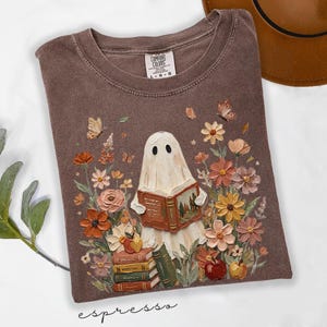 May include: A brown t-shirt featuring a graphic of a white ghost reading an open book, surrounded by colorful flowers, butterflies, and a stack of books with apples. The inner neck label reads Comfort Colors, 100% cotton, size L-G-G, made in Honduras.