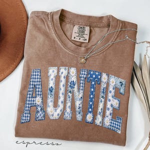 May include: Espresso-colored Comfort Colors t-shirt with the word "AUNTIE" appliqued in blue and white patterned fabric. The shirt features a small tag and a silver necklace with a pendant. The shirt is size S-P-CH.