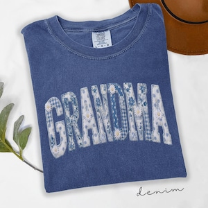 May include: A blue denim t-shirt with the word "GRANDMA" in a patchwork design. The letters are filled with floral and gingham patterns, accented with small white daisies. The shirt has a crew neck and the text "denim" is written in cursive.