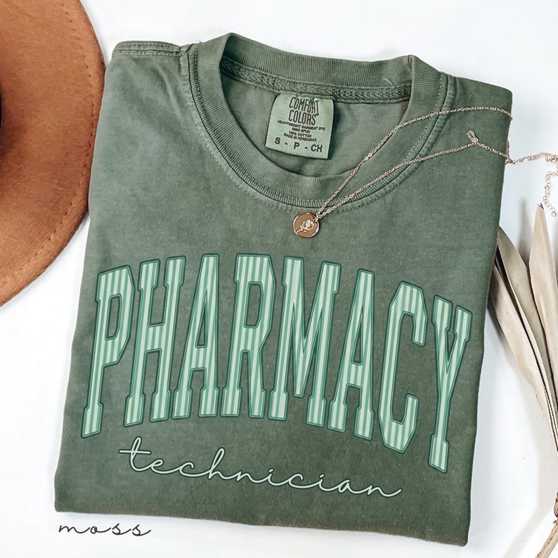 K&ouml;nnte beinhalten: Ein moosgr&uuml;nes Kurzarm-T-Shirt mit dem Wort PHARMACY in gro&szlig;en, hellgr&uuml;n gestreiften Blockbuchstaben und dem darunter in wei&szlig;er Schreibschrift geschriebenen Wort technician. Das T-Shirt hat einen Rundhalsausschnitt und ein Comfort Colors Etikett.