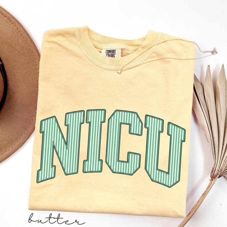 May include: A butter yellow short-sleeve t-shirt with a crew neckline and the text NICU printed in large, green and white vertical striped block letters across the chest. The shirt features a Comfort Colors brand label at the inner neck.