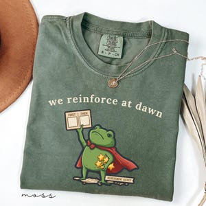 Pode incluir: Uma t-shirt verde-oliva com um gráfico de um sapo usando uma capa vermelha, segurando uma placa com "FIRST THEN" e estrelas amarelas. O texto "we reinforce at dawn" está impresso acima, com "ACHIEVEMENT LOCKED" abaixo do desenho.