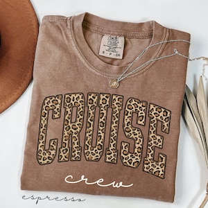 Könnte beinhalten: Braunes T-Shirt mit dem Wort "CRUISE" in Leopardenmuster-Buchstaben. Das Wort "crew" steht darunter in Schreibschrift. Eine silberne Halskette mit Anhänger liegt über dem Shirt. Das Shirt ist "espresso" gefärbt.
