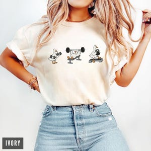 May include: Ivory-coloured t-shirt with a cartoon goose lifting weights. The goose is shown in three poses, using dumbbells, a barbell, and a kettlebell. The word "IVORY" is printed in a black rectangle at the bottom of the shirt.