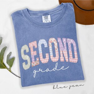 May include: A blue short-sleeve t-shirt with a crew neckline. The front features the text SECOND in large block letters with a multi-colored floral and gingham pattern, and the word grade in white cursive script below the block letters.