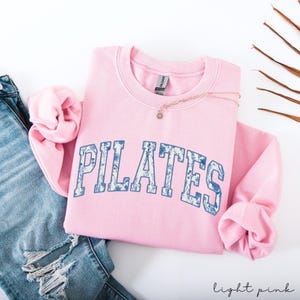 May include: A light pink Pilates sweatshirt with blue floral lettering. The sweatshirt is folded with the sleeves tucked in. A delicate necklace with a small pendant rests on the sweatshirt. The words "light pink" are written in cursive in the bottom right corner.