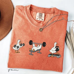 May include: Orange Comfort Colors t-shirt featuring a graphic of three ducks exercising with weights. The ducks are shown lifting dumbbells, bench pressing, and using a kettlebell. A silver necklace with a small pendant is also visible.
