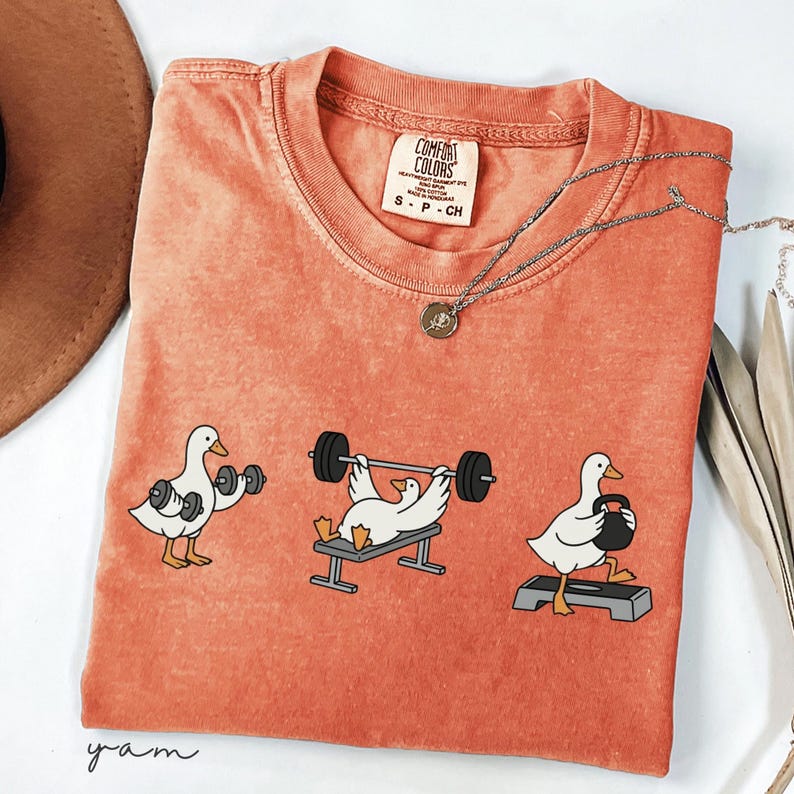 May include: Orange Comfort Colors t-shirt featuring a graphic of three ducks exercising with weights. The ducks are shown lifting dumbbells, bench pressing, and using a kettlebell. A silver necklace with a small pendant is also visible.