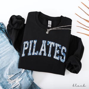 May include: Black crewneck sweatshirt with the word "PILATES" in a blue floral design. A silver necklace with a circular pendant rests on the sweatshirt. The word "black" is written in a script font at the bottom right.