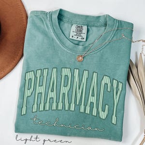 K&ouml;nnte beinhalten: Ein hellgr&uuml;nes Kurzarm-T-Shirt mit Rundhalsausschnitt. Auf der Vorderseite steht das Wort PHARMACY in gro&szlig;en, gebogenen, gestreiften Blockbuchstaben, darunter das Wort technician in Schreibschrift. Eine kleine silberne Halskette liegt darauf.