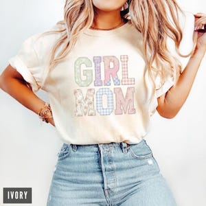 May include: Ivory-colored t-shirt with the words "GIRL MOM" in pastel floral and plaid patterns. The shirt is a relaxed fit and is paired with blue denim jeans. The word "IVORY" is displayed in a black rectangle at the bottom left.