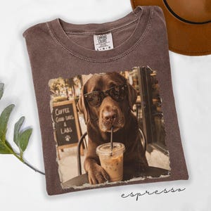 May include: A brown t-shirt featuring a graphic of a chocolate Labrador dog wearing sunglasses and holding a plastic cup with an iced coffee. The graphic includes a sign in the background that reads Coffee Good Days & Labs with a paw print.