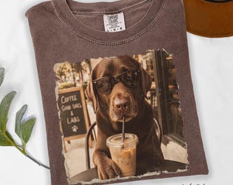 May include: A brown t-shirt featuring a graphic of a chocolate Labrador dog wearing sunglasses and holding a plastic cup with an iced coffee. The graphic includes a sign in the background that reads Coffee Good Days & Labs with a paw print.