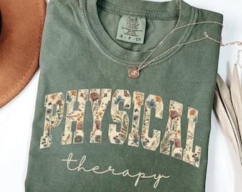 May include: A moss green t-shirt with the words "PHYSICAL THERAPY" in a floral design. The words "therapy" is written in a cursive font. A silver necklace with a pendant is draped across the shirt. The shirt is folded on a white surface.