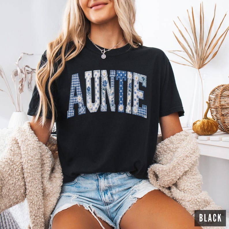 May include: Black t-shirt with the word "AUNTIE" in a patchwork design of blue floral and gingham patterns. The shirt is a crew neck style and is paired with denim shorts. The image is well-lit, showcasing the details of the shirt.