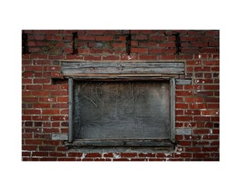 May include: A weathered, boarded-up window set into a red brick wall. The window is framed with rough-hewn wood and covered with a wire mesh. The brickwork shows signs of age and weathering, with variations in color and texture.