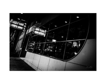 May include: Black and white photograph of a restaurant exterior at night. The building has large windows reflecting the interior. Neon signs read "FOOD & DRINK" and "NOW OPEN". The image is taken at a slight angle.
