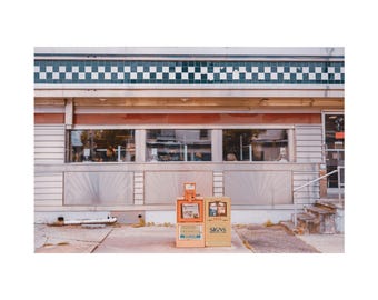 May include: Exterior shot of a retro diner with a red and white facade, featuring large windows and a checkered tile border. Two newspaper stands are in front, one orange and one yellow, with the word "SIGNS" visible.