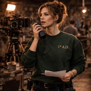 Peut inclure: Une femme portant un sweat-shirt vert foncé avec le texte "1st A.D." en lettres dorées, tenant un talkie-walkie et un script. Elle porte une montre et a un écouteur. Un plateau de tournage est en arrière-plan.