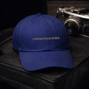 Peut inclure: Casquette de baseball bleu marine avec le mot "CINEMATOGRAPHER" brodé en fil doré. La casquette est posée sur une surface noire, avec un appareil photo vintage en arrière-plan.