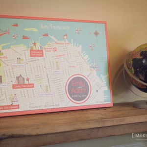 May include: A red and white illustrated map of San Francisco with the text "Emily and Adam June 14, 2014" in a circle on the map. A globe is on a wooden table in the background.