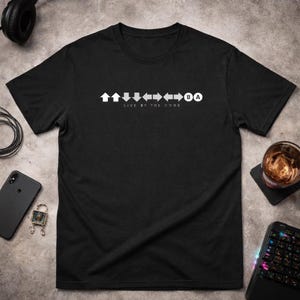 Könnte beinhalten: Schwarzes T-Shirt mit einer weißen Grafik aus Pfeilen und dem Text "LIVE BY THE CODE". Das Design umfasst Pfeile nach oben, unten, links und rechts sowie die Tasten "A" und "B". Ein modernes Design für Gamer.