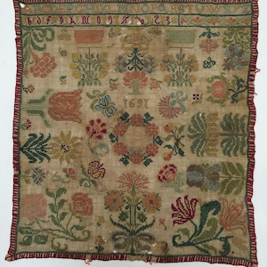 May include: A square needlepoint sampler on beige fabric featuring a red striped border. The design includes an alphabet, the year 1691, and various floral motifs in green, red, yellow, and blue thread. The embroidery displays symmetrical plant patterns.