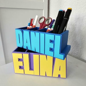 Könnte beinhalten: Ein zweistufiger Schreibtisch-Organizer in Blau, Gelb und Lila, mit den Namen "DANIEL" und "ELINA" in großen Blockbuchstaben. Der obere Bereich enthält Stifte, Marker, Scheren und Klebestifte. Der Organizer ist aus Kunststoff.