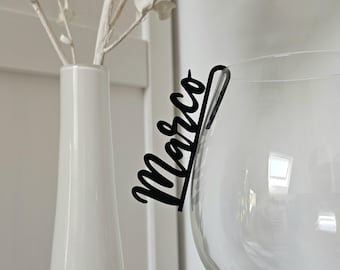 May include: A black acrylic wine glass charm featuring the name Marco in a cursive script font. The charm is designed to hook onto the rim of a clear glass, providing a personalized identifier for the drinkware during social gatherings.