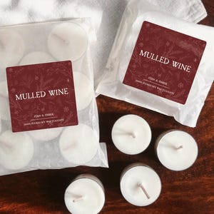 May include: Two packages of white soy wax tealights with a burgundy label that reads "MULLED WINE". The labels also include the text "FERN & EMBER" and "HAND POURED SOY WAX TEALIGHTS". Several loose tealights are scattered around the packages.