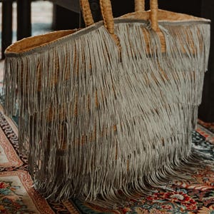 Puede incluir: Un bolso tote de paja tejido con un ribete de flecos. El bolso tiene dos asas tejidas y es de color marrón claro con flecos plateados.