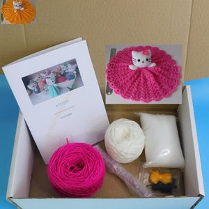 May include: A craft kit for making a kitten doll. The kit includes pink and white yarn, stuffing, a crochet pattern booklet, and a small, finished kitten doll. The finished doll is white with a pink bow and a pink crocheted skirt.