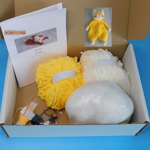 May include: A craft kit in a cardboard box, featuring a yellow and white yarn, a small finished yellow cat toy, and a pattern titled "Kitty." The kit includes various small craft supplies and a white, fluffy filling.