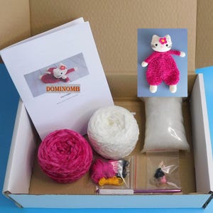 May include: A craft kit containing pink and white yarn, a crochet hook, and other supplies. The kit includes a pattern and a finished pink and white crocheted cat. The text "DOMINOMB" is visible on the pattern.