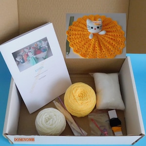 May include: A craft kit for making a kitten doll, presented in a cardboard box. The kit includes yarn in yellow and white, a white pillow, a crochet hook, and a pattern booklet. A finished kitten doll with a yellow crocheted skirt is also included.