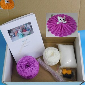 May include: A craft kit in a cardboard box, featuring a kitten-themed crochet project. The kit includes purple and white yarn, stuffing, a crochet hook, and a pattern booklet. A finished kitten doll is shown in the image.