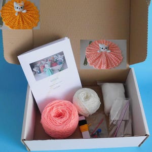 May include: A craft kit for making kitten dolls, presented in an open cardboard box. The kit includes yarn in pink and white, a pattern booklet, and other supplies. Two finished kitten dolls are shown, one in orange and one in pink.