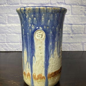 May include: A cylindrical ceramic tumbler featuring a textured, off-white base glaze with vibrant blue glaze dripping from the rim. A circular stamp with the letter S is embossed near the top. A thin, horizontal band of brown glaze accents the lower section.