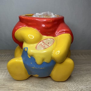 May include: A vintage Winnie the Pooh ceramic jar in the shape of the character. The jar is primarily yellow with red and blue accents. The jar has a label that reads "Winnie the Pooh Collection".