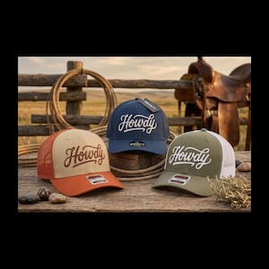 May include: Three baseball caps with the word "Howdy" in white script. The caps are in different colours: orange and tan, navy blue, and olive green. The caps are on a wooden surface with a rope and a saddle in the background.