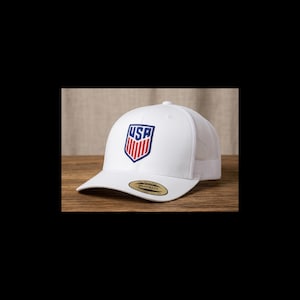 May include: White baseball cap with a mesh back and a curved brim. The front of the cap features a shield-shaped emblem with the letters "USA" in blue and red. The cap has a snapback closure.