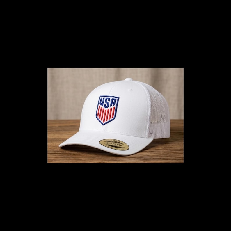 May include: White baseball cap with a mesh back and a curved brim. The front of the cap features a shield-shaped emblem with the letters "USA" in blue and red. The cap has a snapback closure.