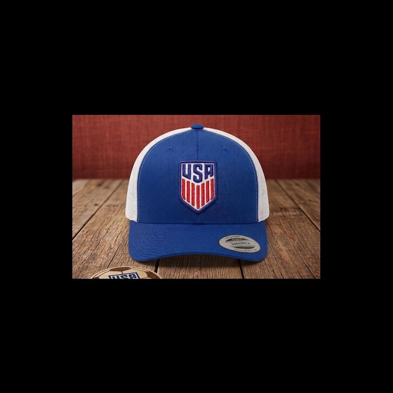 May include: A blue and white baseball cap with the word "USA" in a shield design on the front. The cap has a curved brim and a mesh back. The hat is displayed on a wooden surface.