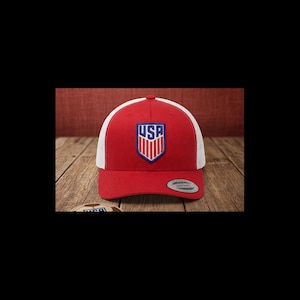 May include: A red and white baseball cap with a USA logo on the front. The cap has a curved brim and a mesh back. The USA logo is in a shield shape with red, white, and blue colors.